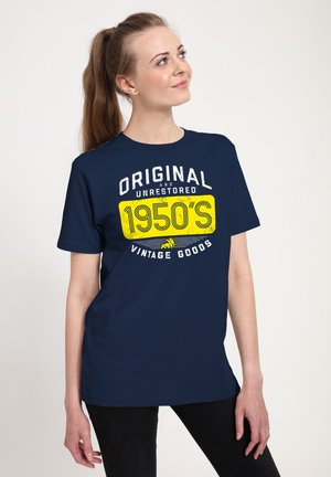 Młoda kobieta z kitką, ubraną w granatowy T-shirt z napisem „Original and Unrestored 1950's Vintage Goods”, spoglądająca w górę i na bok.