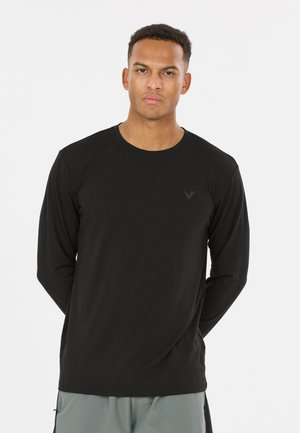 Jeune homme aux cheveux courts portant une chemise noire à manches longues et un pantalon gris, debout les mains dans le dos sur un fond blanc.