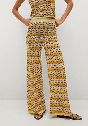 Kvinde iført højtaljede vidde bukser med gul, beige og brun zigzag-mønster, kombineret med matchende ærmeløs top og sandaler.