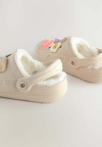 Chaussures beiges avec une doublure en fourrure douce, ornées de patches décoratifs, y compris des icônes alimentaires. Design à l'arrière fermé avec un détail de bouton sur le côté.