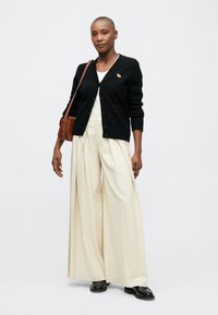 Schwarze gestrickte Strickjacke mit einer kleinen dekorativen Nadel, kombiniert mit lockeren, gefalteten elfenbeinfarbenen Hosen und schwarzen Schuhen, was einen kontrastierenden Look schafft.