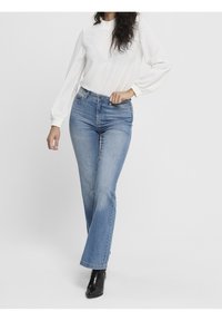 Nainen, joka pukeutuu valkoiseen pitkähiharaiseen paitaan, vaaleansinisiin korkeavyötäröisiin farkkuihin ja mustiin nilkkabootseihin, seisoo käsivarsi lantiolla.