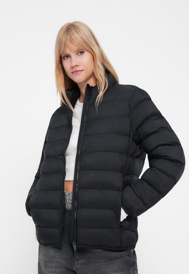 Chaqueta acolchada negra con acolchado horizontal, cuello alto, cremallera frontal y bolsillos laterales. La lleva puesta sobre una parte superior blanca recortada y pantalones grises.