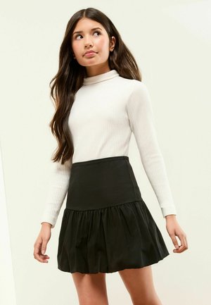 Tânără cu păr lung și închis la culoare, purtând un top alb cu mâneci lungi și țesătură ribată și o fustă mini neagră cu pliuri, privind în lateral.