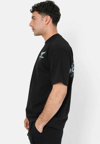 Jonge man met kort, krullend haar, gekleed in een zwart grafisch t-shirt en zwarte broek, die in profiel staat met zijn handen in zijn zakken.