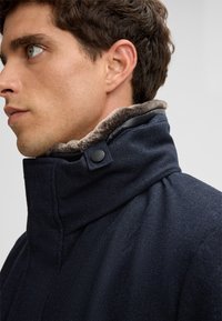 Cappotto di lana blu scuro con colletto alto, dotato di una fodera in pelliccia sintetica grigia e un bottone decorativo nero. Il tessuto ha una finitura textured.