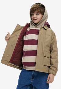 Tan ytterjacka med dragkedja och tryckknappar, med ett maroon-fodrat inre. Under jackan en randig burgundy och krämfärgad hoodie. Blå jeans.