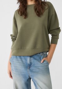 Olijfgroene sweatshirt met verlaagde schouders en manchet mouwen, gecombineerd met lichtblauwe high-waisted jeans. Zachte stof textuur.