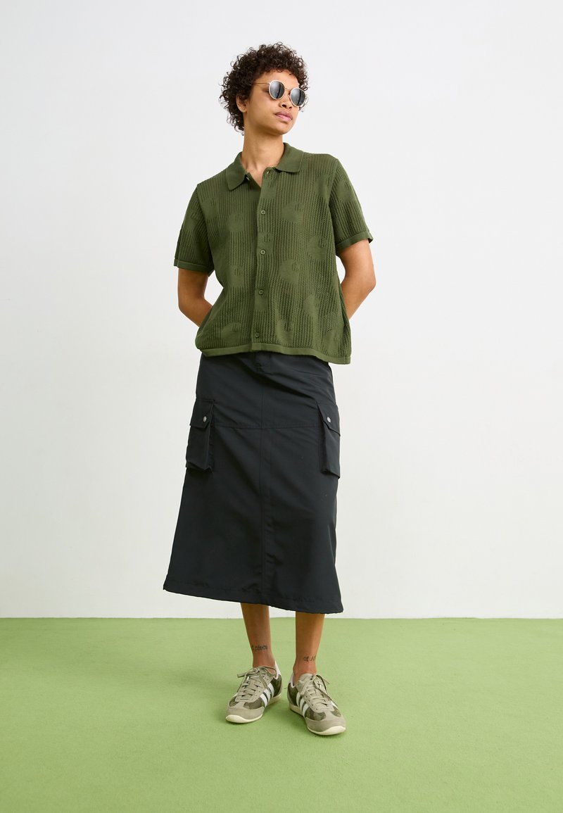 Olijfgroene polo met korte mouwen en een gestructureerd patroon, gecombineerd met een zwarte rok tot de knie met cargopockets. Neutrale sneakers.