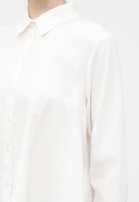 Κοντινό πλάνο ενός ατόμου που φοράει ένα απλό λευκό μακρυμάνικο πουκάμισο με κουμπιά και κλασικό γιακά, μπροστά σε ουδέτερο φόντο.