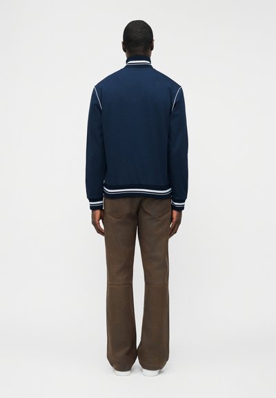 Homme debout, tourné de dos, portant une veste bleu marine avec un liseré blanc et un pantalon large marron avec des baskets blanches.