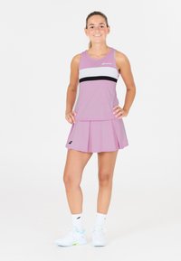 Giovane donna in piedi che indossa un completo da tennis rosa e bianco, con sneakers e calze bianche, sorridente con le mani sui fianchi su uno sfondo bianco.