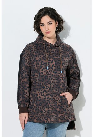 Kvinde med kort, mørkt hår iført øreringe med ringe og en brun leopardmønstret hættetrøje med forlomme og lyseblå jeans.