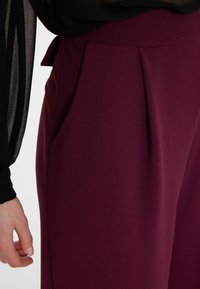 Bordeaux hoogtaille broeken met een gladde textuur, voorzien van plooien en zijzakken. De tailleband is versierd met een klein strikje.