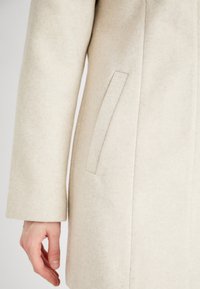 Beige ullkappa med en strukturerad design, med en sidoficka och en slät yta, visad från armbågen neråt med en hand vilande.