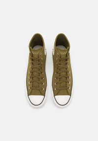Green canvas high-top sneakers with white rubber toe caps, round laces, and a circular logo on the outer sides. Textured upper with stitching details.
