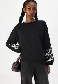 Svart genser med lange ermer med dekorativ hvit brodering. Kombinert med mørke jeans og en liten brun lær crossbody-veske.