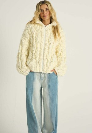 Kobieta o długich blond włosach, ubrana w kremowy, gruby sweter rozpinany na suwak z grubym splotem oraz dżinsy z szerokimi nogawkami w dwóch odcieniach, z rękami w kieszeniach.