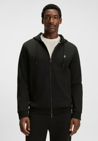 Uomo che indossa una felpa nera con zip e cappuccio a strisce sopra una maglietta bianca, in piedi contro uno sfondo chiaro e uniforme.