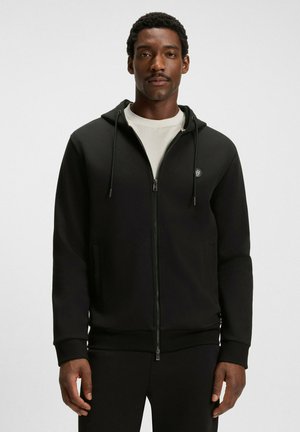 Uomo che indossa una felpa nera con zip e cappuccio a strisce sopra una maglietta bianca, in piedi contro uno sfondo chiaro e uniforme.