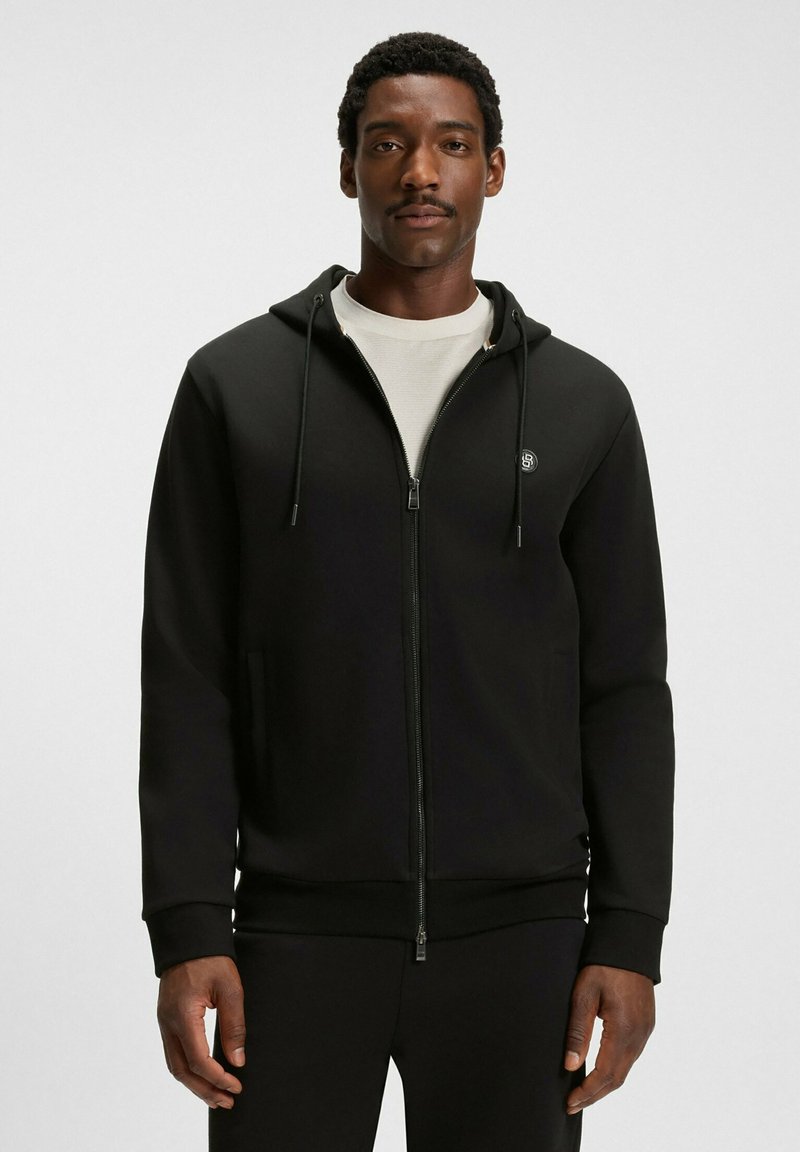 Uomo che indossa una felpa nera con zip e cappuccio a strisce sopra una maglietta bianca, in piedi contro uno sfondo chiaro e uniforme.