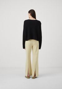 Svart kort cardigan med teksturert finish, kombinert med lysegule høytaljede bukser med splitt i kanten. Uformelt fottøy synlig.