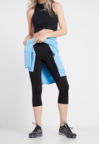 Femme portant un legging noir court et un crop top sans manches, avec un sweat-shirt bleu noué autour de la taille, debout sur fond blanc.