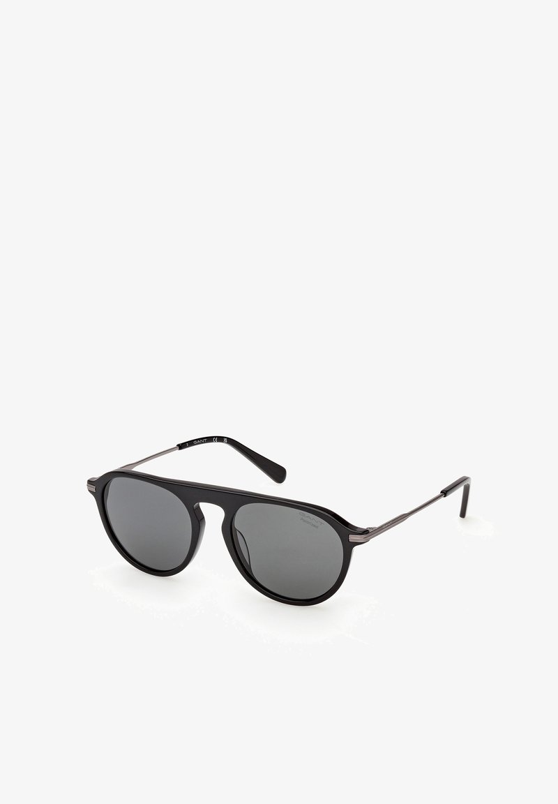 Lunettes de soleil de style aviateur noires avec des verres foncés et des branches fines en métal, inclinées sur un fond blanc.
