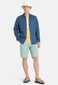 Camisa azul de botones sobre una camiseta gráfica amarilla, pantalones cortos verde menta y sandalias multicolores. Lleva un sombrero de cubo blanco.