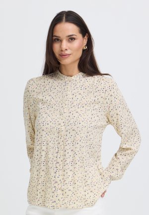 Mujer con cabello largo y oscuro que lleva una blusa color crema con un pequeño estampado floral, de pie con una mano en la cadera frente a un fondo liso.