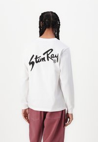 Long-sleeve white T-shirt featuring a large black "Stan Ray" logo on the back. The shirt is paired with loose-fitting pink pants.
