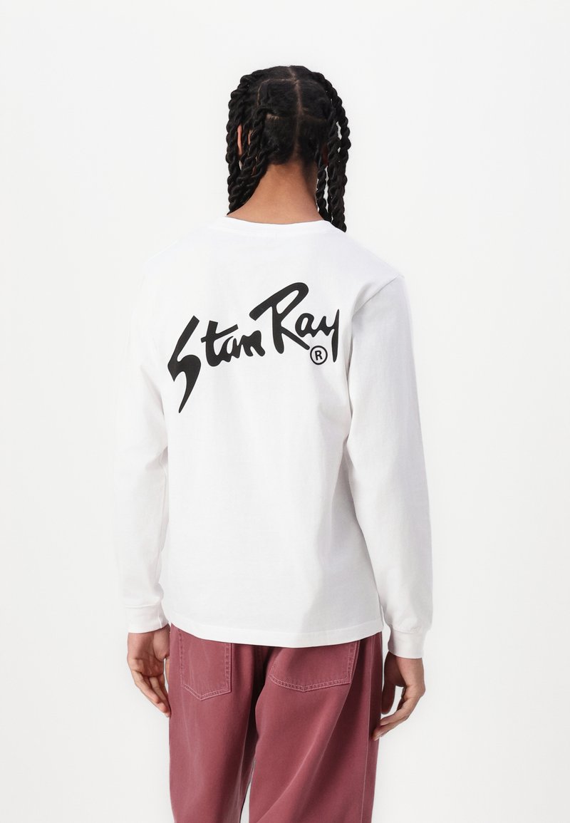 Long-sleeve white T-shirt featuring a large black "Stan Ray" logo on the back. The shirt is paired with loose-fitting pink pants.