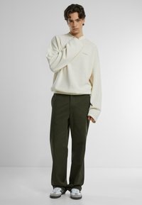 Cremesfarbener, gerippter Strickpullover mit übergroßen Ärmeln, kombiniert mit dunkelgrünen, weit geschnittenen Hosen und hellgrauen Sneakers. Einfaches Design, minimale Akzente.