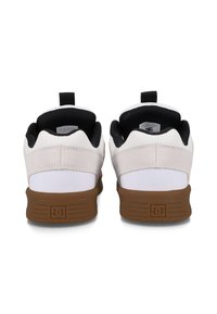 Baskets blanches et noires avec un dessus texturé, équipées d'une semelle en caoutchouc marron et d'une languette à l'arrière pour un enfilage facile. Logo DC sur le talon.