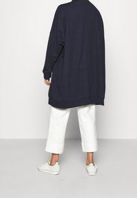 Übergroßer dunkelblauer Kapuzenpullover, weicher Stoff, überschnittene Schultern, Seitenschlitze. Kombiniert mit cropped weißen Hosen und hellen Sneakern.