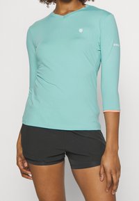 Türkises Langarmoberteil mit V-Ausschnitt, sportlichem Design, kleinem Logo auf der Brust und kontrastierenden orangefarbenen Akzenten an den Ärmeln.