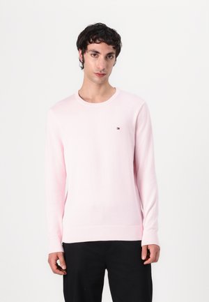 Młody mężczyzna o ciemnych kręconych włosach, ubrany w jasnoróżowy sweter z długim rękawem i czarne spodnie, stojący na jednolitym białym tle.