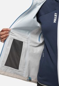 Marineblaue Softshell-Jacke mit einer hellgrauen Fütterung, ausgestattet mit einer Mesh-Tasche und einer Kordelzugverstellung am Saum. Mit durchgehendem Reißverschluss vorne.