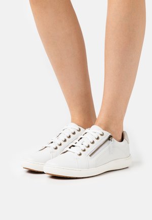 Pieds portant des baskets blanches basses avec lacets et fermoir zippé latéral, posés sur une surface blanche.