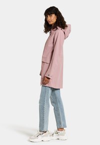 Waterdichte roze regenjas met capuchon, frontzakken en een losse pasvorm, gecombineerd met lichtblauwe jeans en neutrale sneakers.