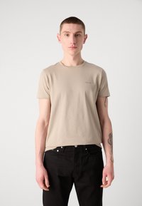 Κοντομάνικη, μπεζ μπλούζα από απαλό ύφασμα, με στρογγυλή λαιμόκοψη και μια διακριτική λεπτομέρεια λογότυπου στο στήθος, σε συνδυασμό με μαύρο παντελόνι.