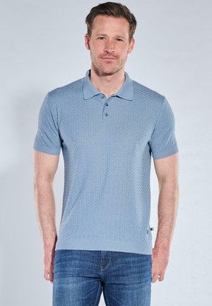 Man standing, wearing light blue textured short-sleeve polo shirt and blue jeans, neutral expression, plain light background.