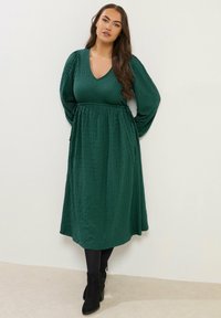 Långärmad, v-ringad, mörkgrön klänning med texturerat mönster, åtsittande midja och en svängd kjol, ihopparad med svarta ankelstövlar.