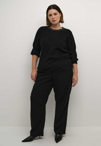 Zwarte gebreide trui met driekwartmouwen en een ronde hals, gecombineerd met losse zwarte broek en zwarte puntige hakken.