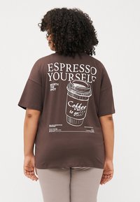 Cilvēks ar cirtainiem matiem valkā brūnu T-kreklu ar kafijas krūzes attēliem un tekstu "ESPRESSO YOURSELF." Viņš ir vērsts prom no kameras.