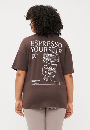 Cilvēks ar cirtainiem matiem valkā brūnu T-kreklu ar kafijas krūzes attēliem un tekstu "ESPRESSO YOURSELF." Viņš ir vērsts prom no kameras.