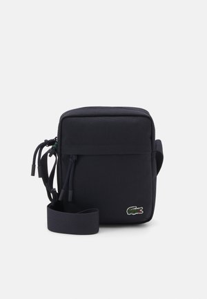 Marineblå stof crossbody taske med lynlås, justerbar rem og et lille grønt broderet krokodillelogo på forsiden.