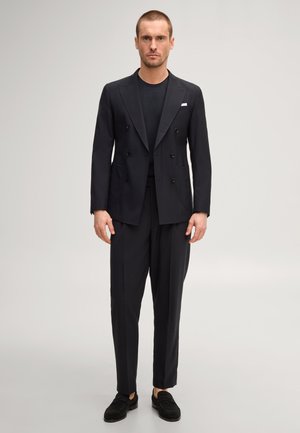 Homme debout portant un blazer noir croisé, une chemise noire, un pantalon noir taillé sur mesure et des mocassins noirs sur un fond clair uni.