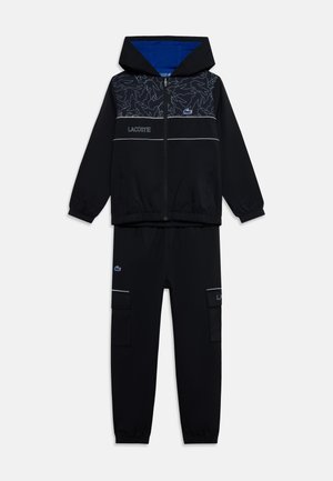 Čierna mikina s kapucňou na zips s abstraktným vzorom a logom Lacoste v kombinácii s čiernymi cargo nohavicami s prednými vreckami a logovými detailmi.