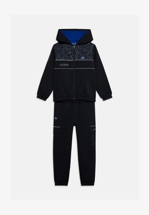 Veste noire à capuche zippée avec motif abstrait et logo Lacoste, associée à un pantalon cargo noir assorti avec poches avant et détails du logo.